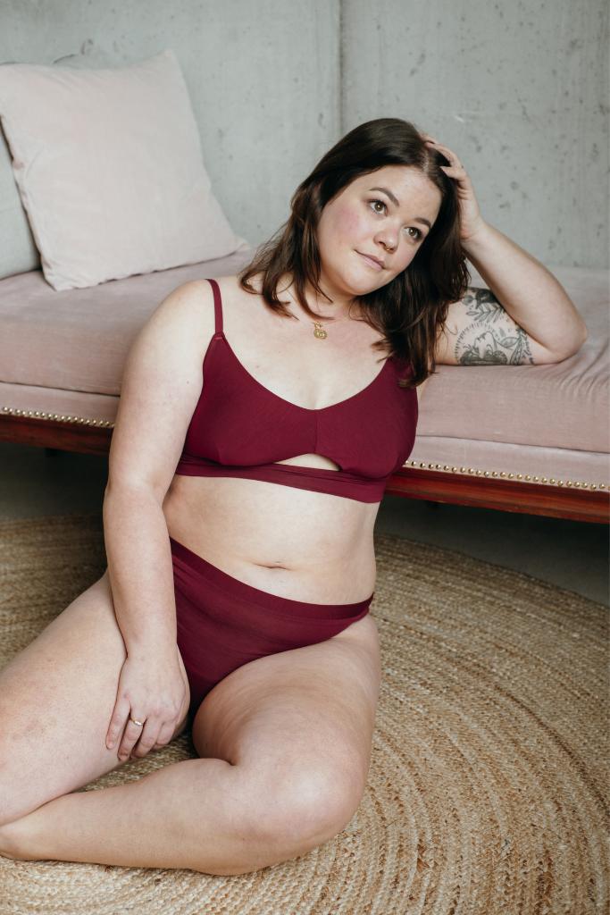 La modella si siede sul pavimento e si appoggia al divano rosa con Thea Bralette e slip rossi.