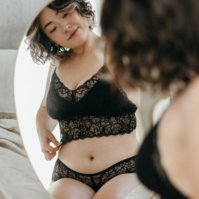 Woman kneels on bed and looks at herself and her black lingerie in the mirror