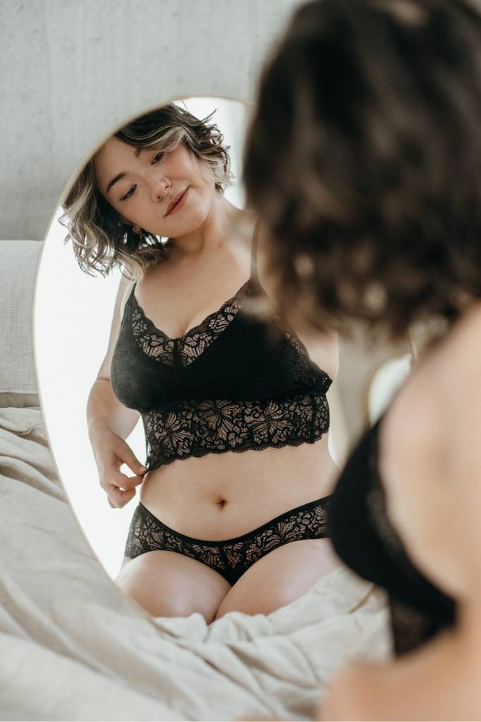 Frau kniet auf Bett und betrachtet sich und ihre schwarze Lingerie im Spiegel