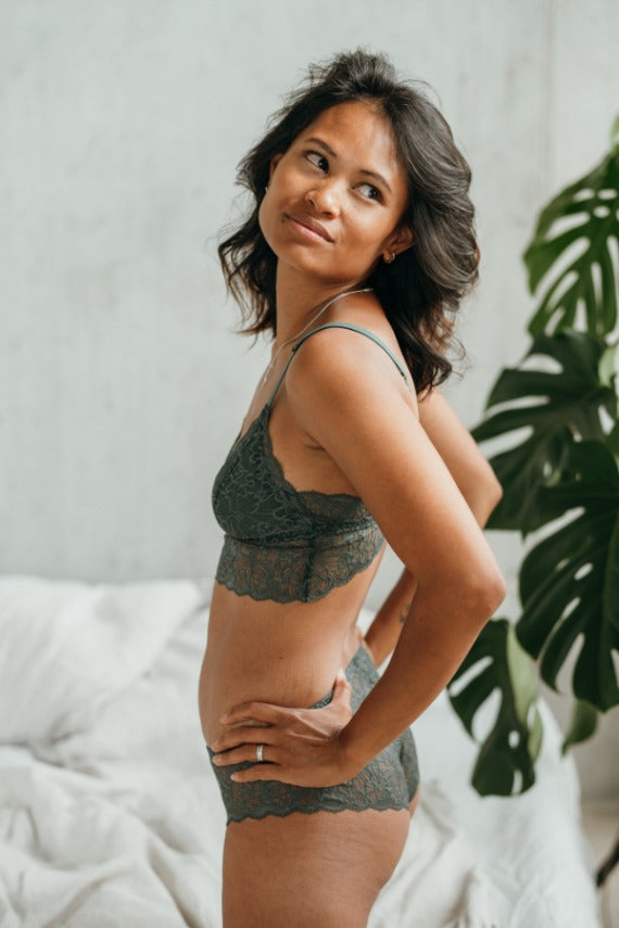 Model in playful lingerie kneels on the side of the bed and looks over her shoulder.