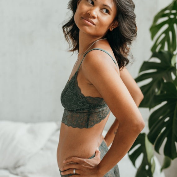 Model in playful lingerie kneels on the side of the bed and looks over her shoulder.