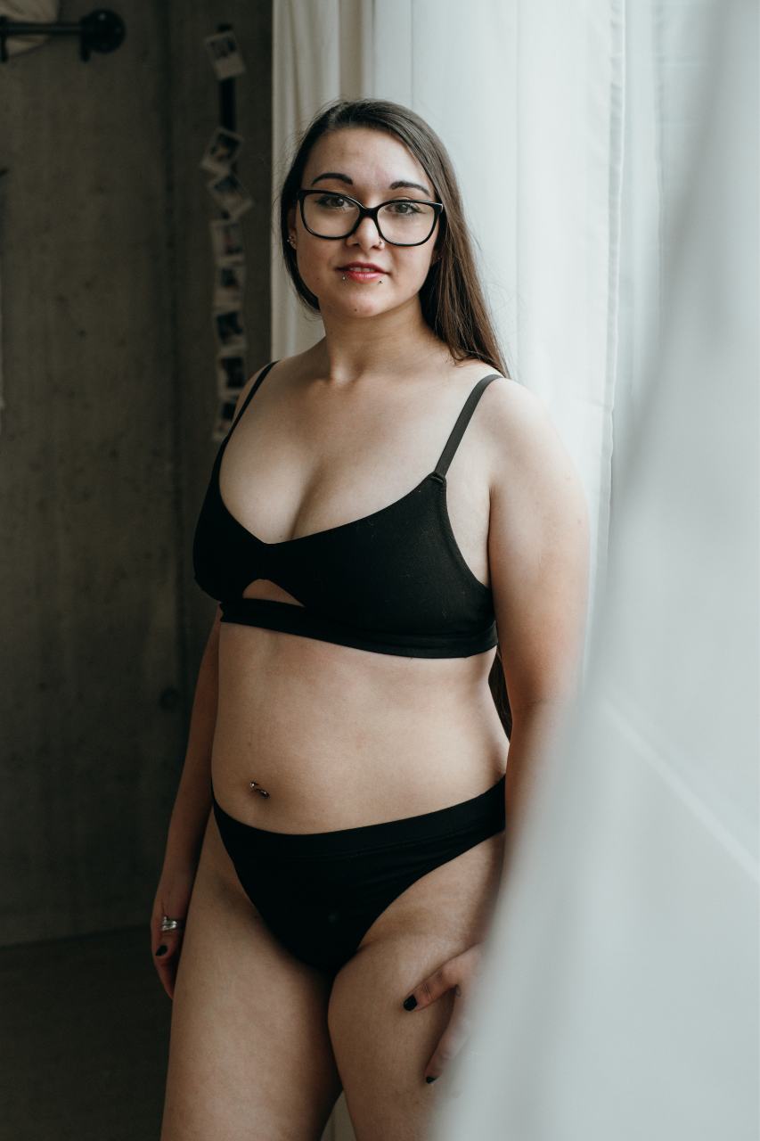 Lors d'un shooting boudoir, le modèle se tient en lingerie noire devant un rideau blanc.