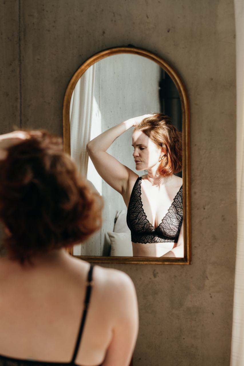 Boudoir photography - model in romantic lingerie looking at herself in the wall mirror.