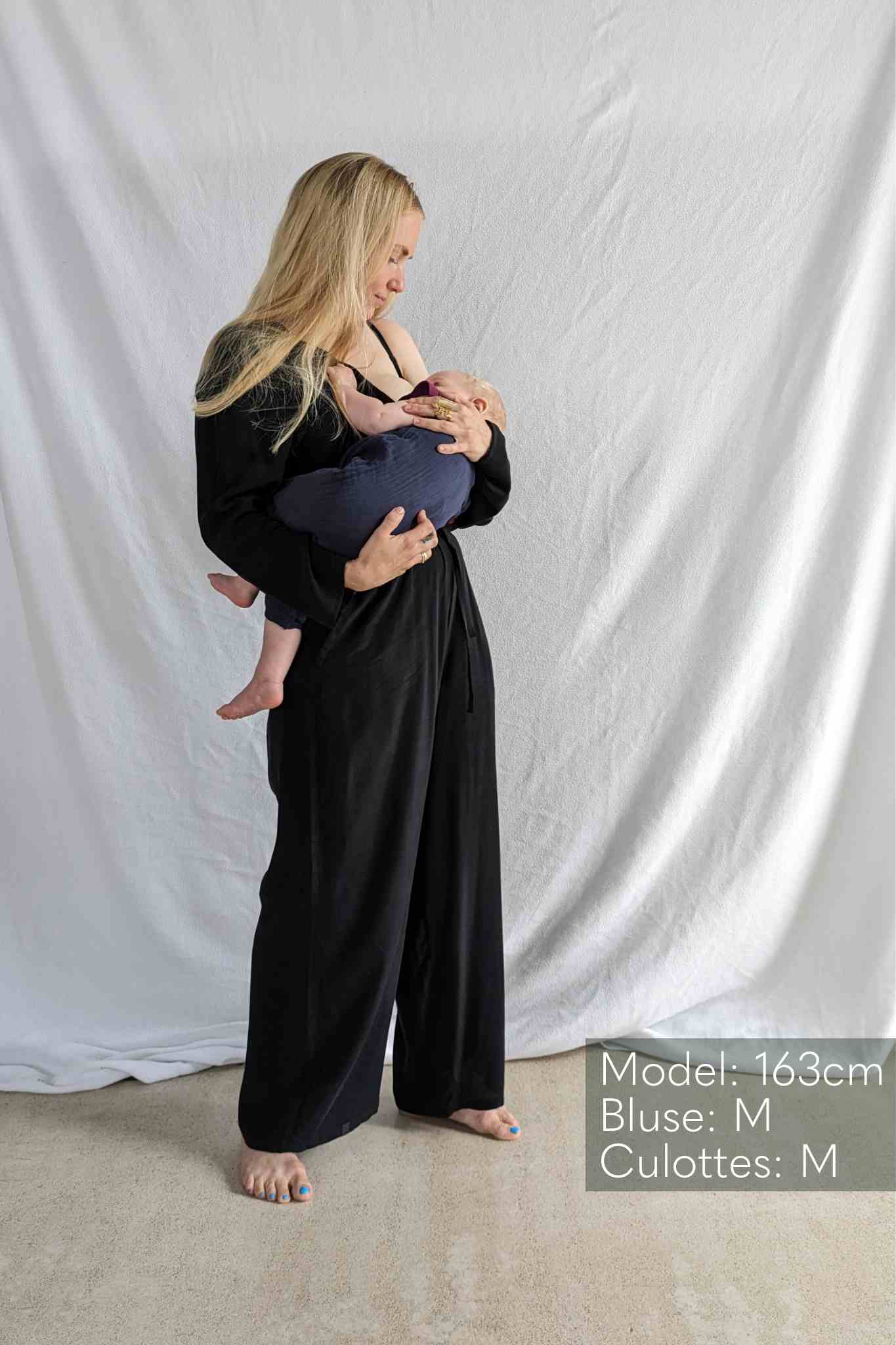 Woman in black blouse and culottes carries baby in her arms.