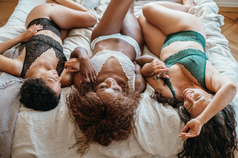 Three different women lie on the bed in their underwear and laugh.