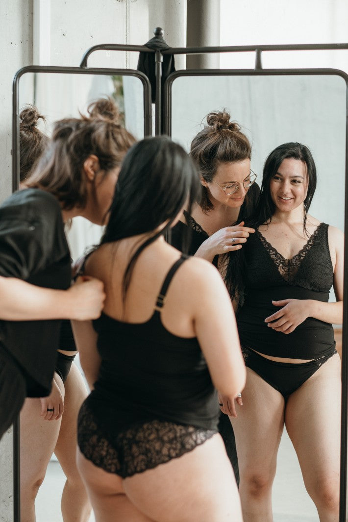 Zippora justiert den Träger eines Nelly Unterhemds an einer Kundin. Sie stehen vor dem Spiegel und lachen.