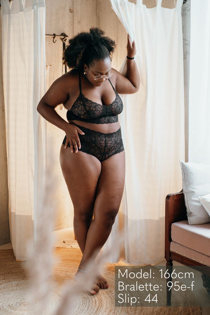 the woman in the black underwear pulls aside the curtain and looks down at herself 
