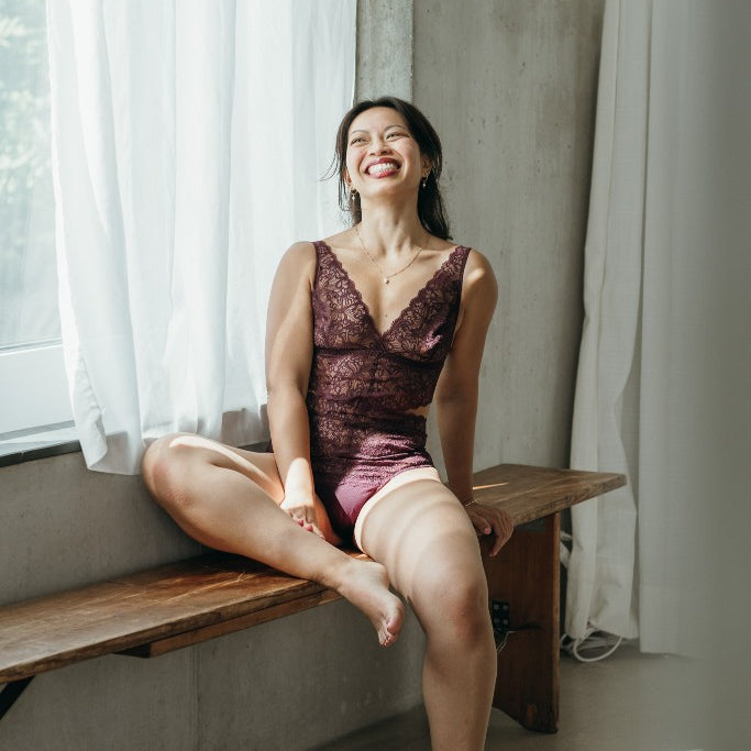 Model sitzt im Alison Body in der Farbe Aubergine auf einer Sitzbank. Ein Bein liegt auf der Bank, sie lacht in die Kamera.