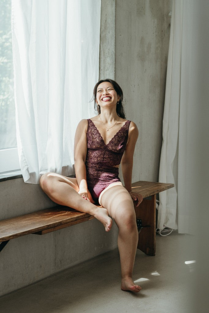 Model sitzt im Alison Body in der Farbe Aubergine auf einer Sitzbank. Ein Bein liegt auf der Bank, sie lacht in die Kamera.