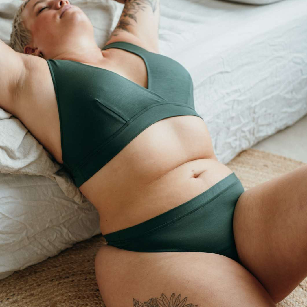 Model trägt nachhaltige Unterwäsche, sitzt vor dem Bett und lehnt sich über die Kante.