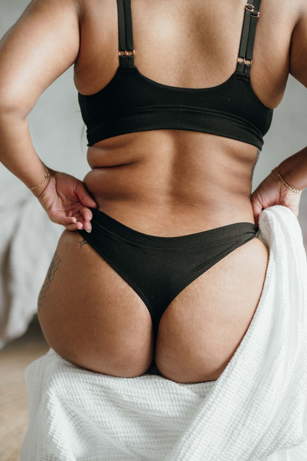 Model sits on chair with blanket. You can see her back, the black String and the Bralette.