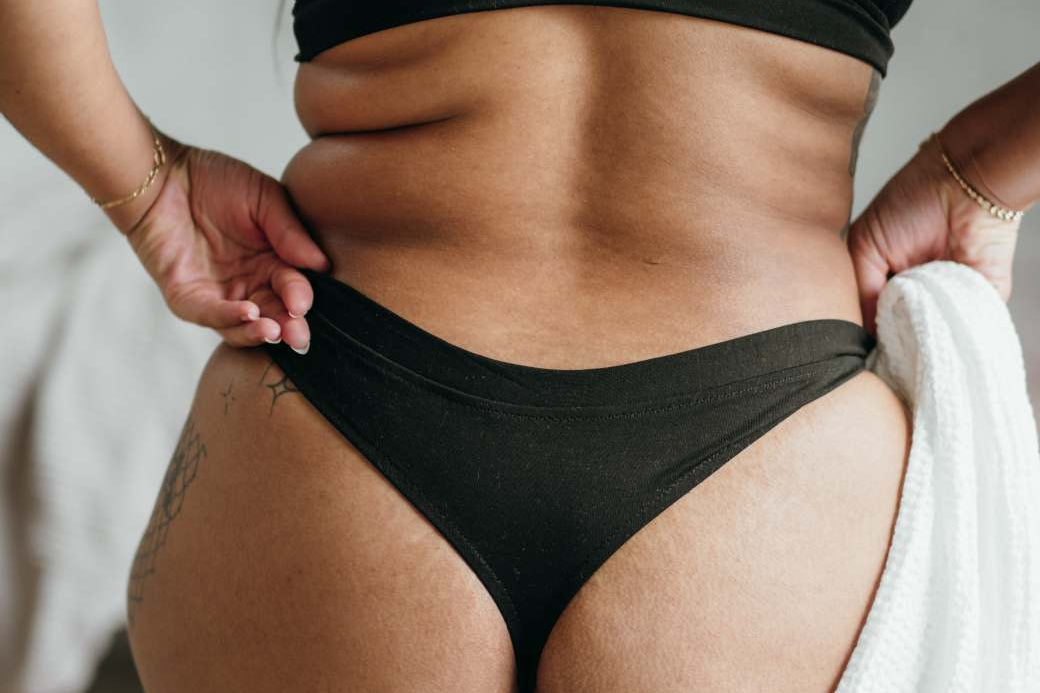 Model sitzt mit Decke auf Stuhl. Man sieht ihren Rücken, den schwarzen String und das Bralette.