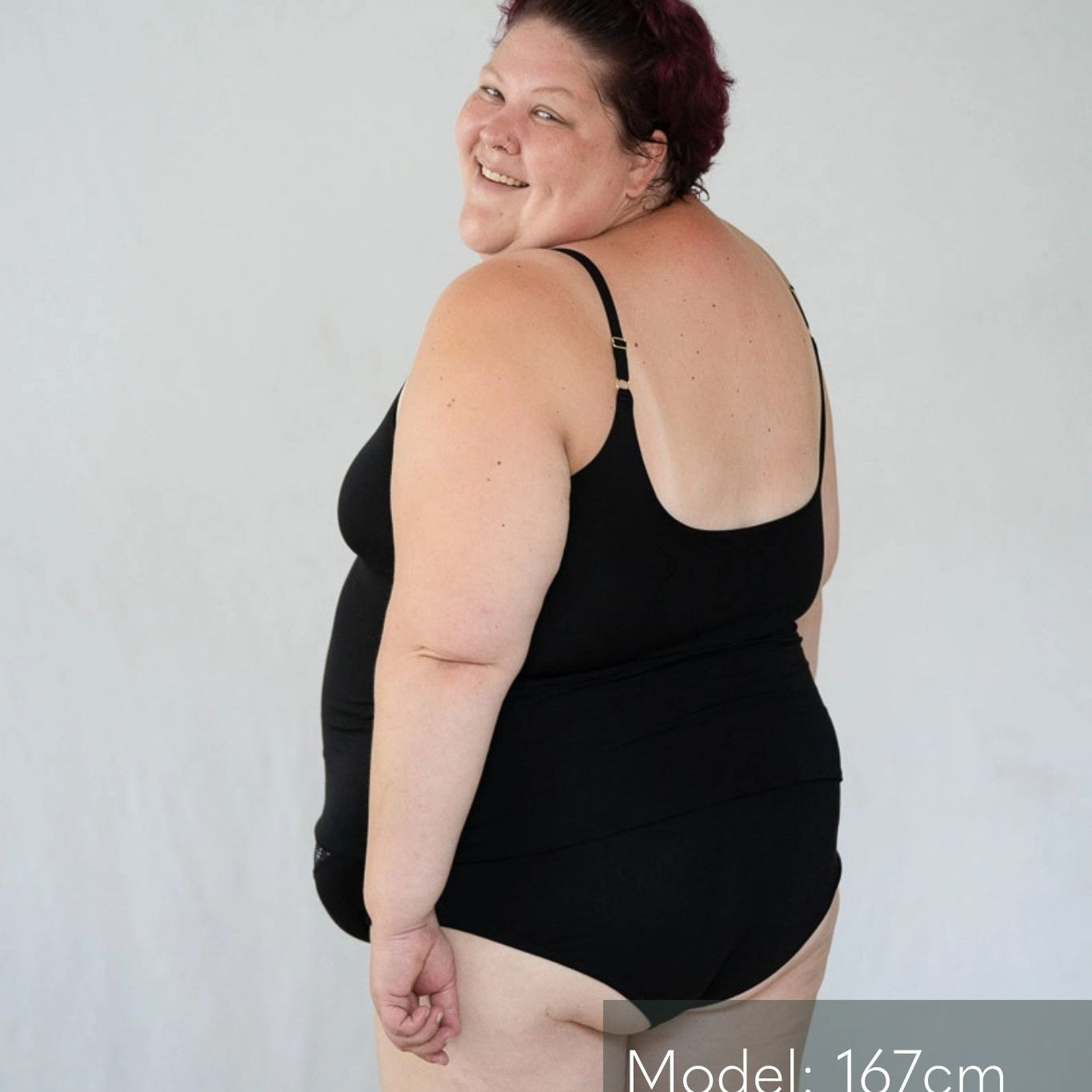 Model steht mit dem Rücken zur Kamera vor weissem Hintergrund und blickt über die Schulter nach hinten. Sie trägt das Carla Camitop und den Nelly Slip in Schwarz.