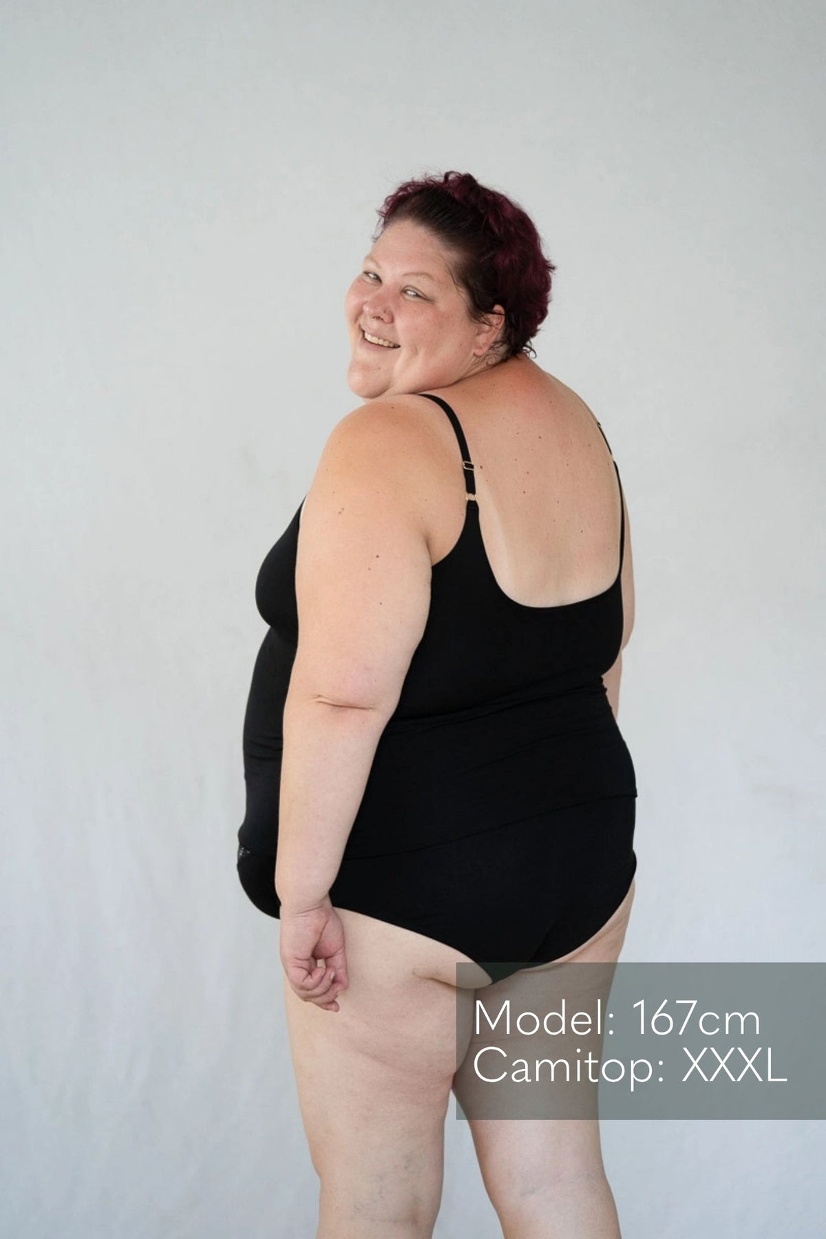 Model steht mit dem Rücken zur Kamera vor weissem Hintergrund und blickt über die Schulter nach hinten. Sie trägt das Carla Camitop und den Nelly Slip in Schwarz.
