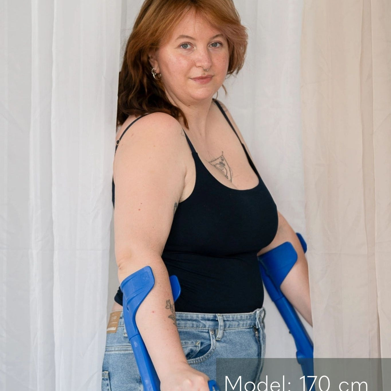 Model steht auf Krücken zwischen Vorhängen und blickt direkt in die Kamera. Sie trägt das Carla Camitop in Night Blue und dazu Jeans Hosen.