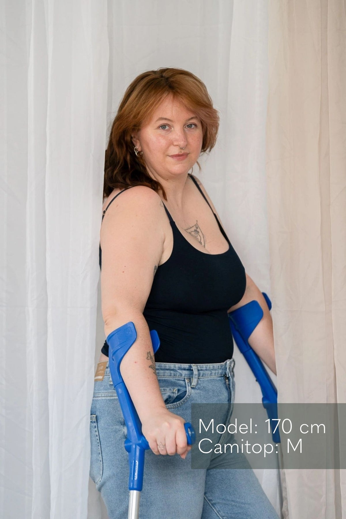 Model steht auf Krücken zwischen Vorhängen und blickt direkt in die Kamera. Sie trägt das Carla Camitop in Night Blue und dazu Jeans Hosen.