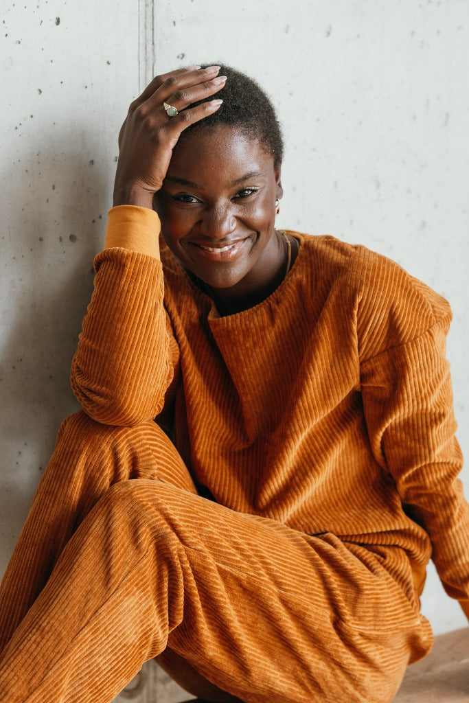 Model trägt ockerfarbiges Pyjama in Cord. Sie fasst sich mit der einen Hand ins kurze Haar und lächelt direkt in die Kamera