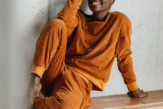 Model trägt ockerfarbigen Pyjama. Sie sitzt auf einer Sitzbank, die Beine auf der Bank angewinkelt und stützt den Kopf auf dem Ellbogen ab.