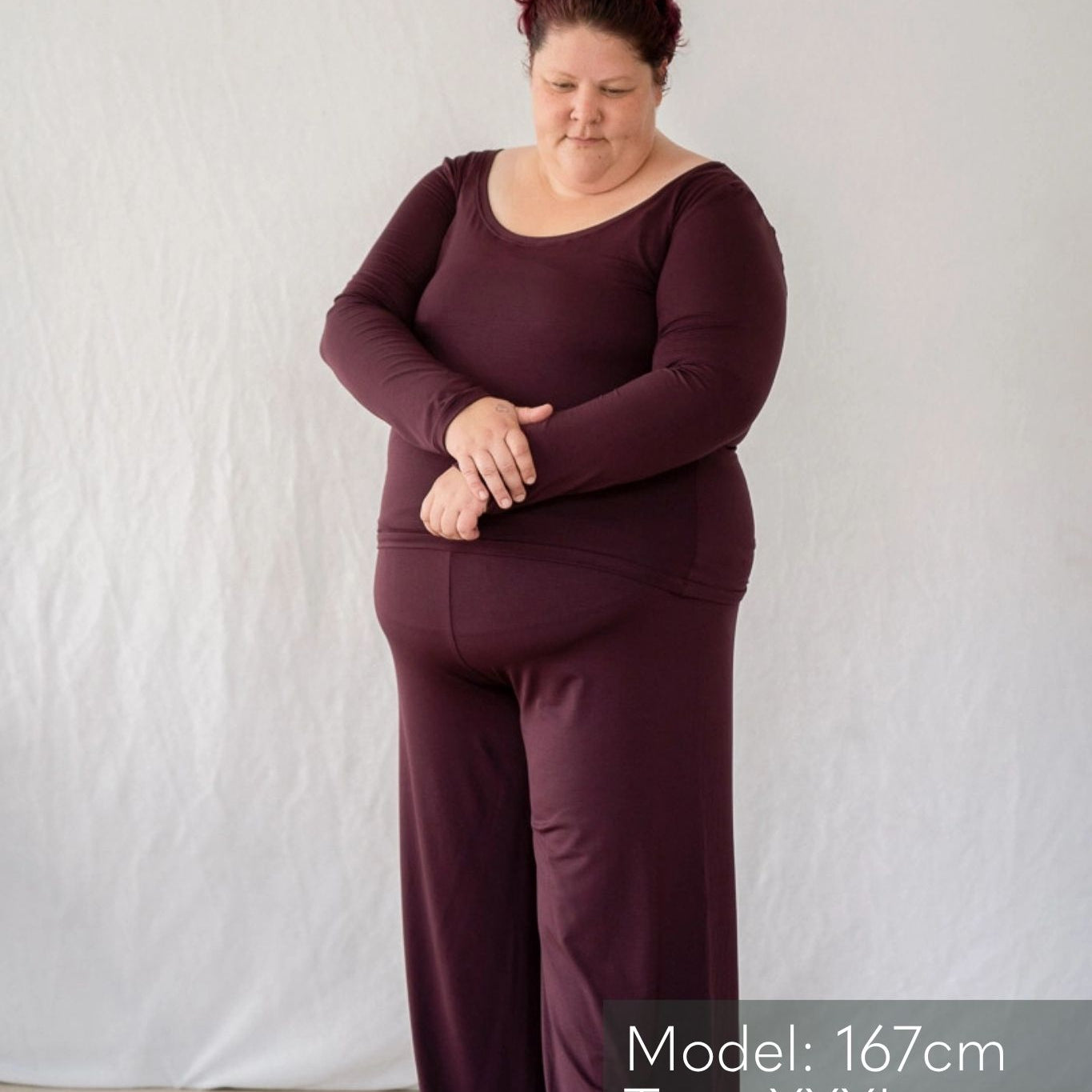 Model steht vor weissem Hintergrund und blickt verträumt zu Boden. Sie trägt das Dolly Longsleeve und die Renée Hose in Aubergine.
