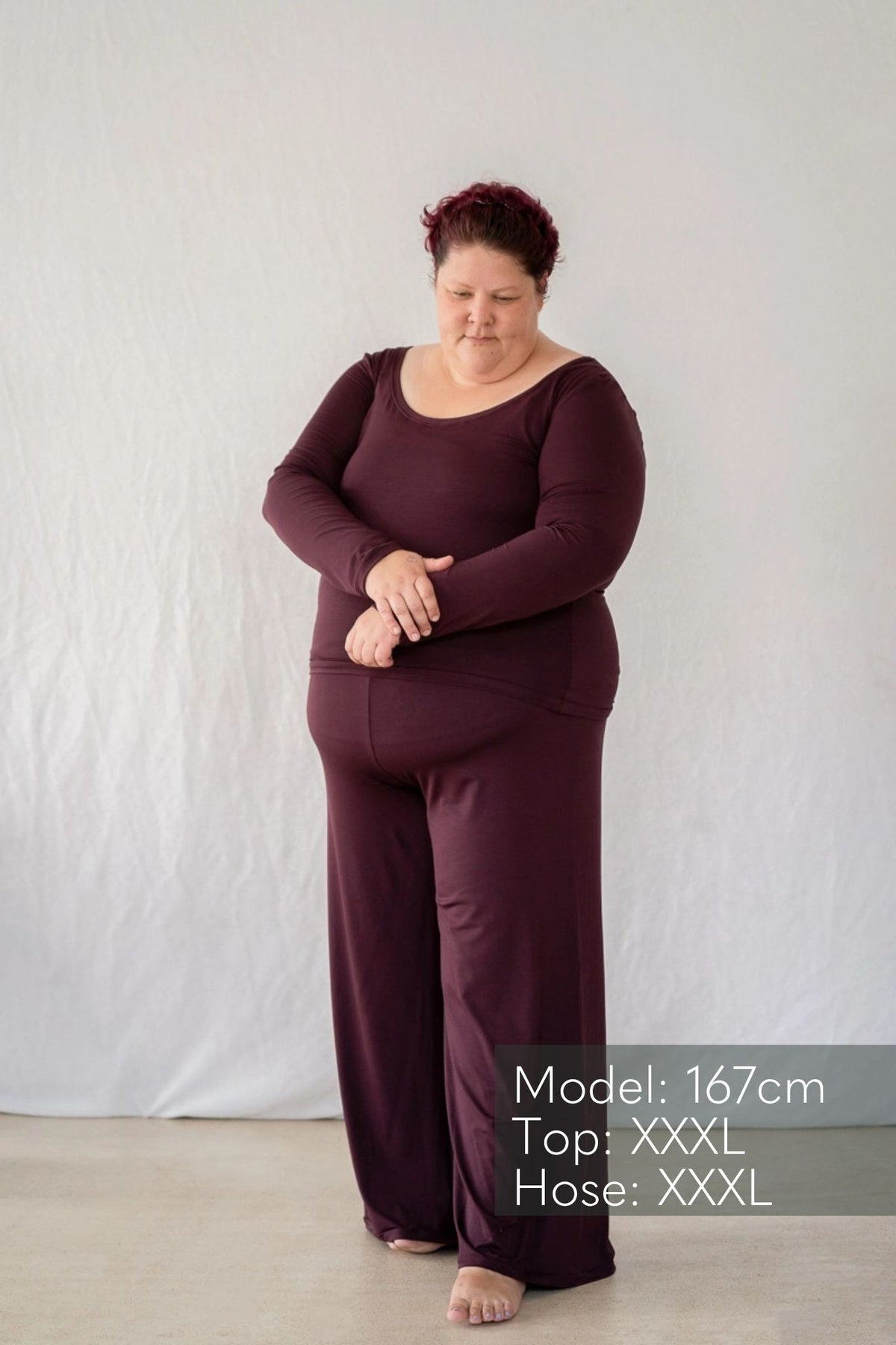 Model steht vor weissem Hintergrund und blickt verträumt zu Boden. Sie trägt das Dolly Longsleeve und die Renée Hose in Aubergine.