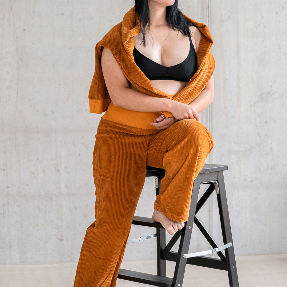 Model trägt Pyjama in ockerfarbigem Cord mit weichen Bündchen, den Pullover hat sie lässig über die Schultern gelegt.