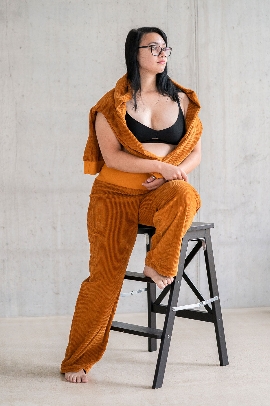 Model trägt Pyjama in ockerfarbigem Cord mit weichen Bündchen, den Pullover hat sie lässig über die Schultern gelegt.