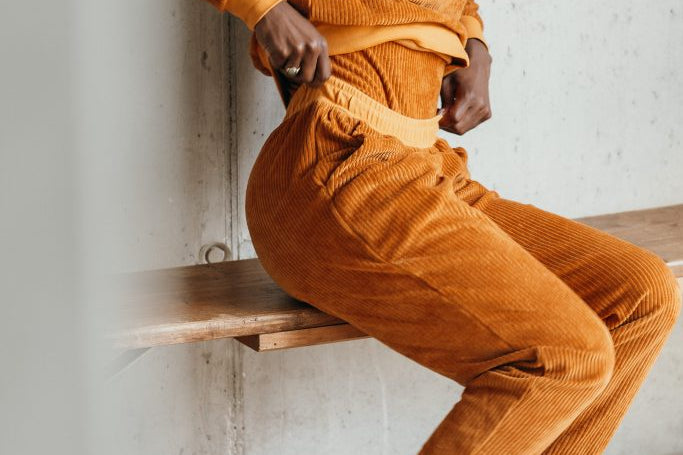 Model trägt den Pyjama aus ockerfarbigem Cord. Sie sitzt auf einer Sitzbank und zupft am Bund