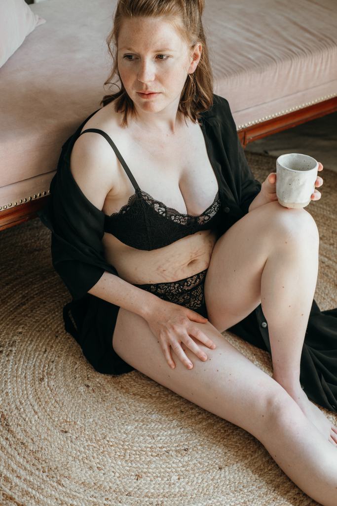 Woman in black lingerie leans against sofa and drinks coffee