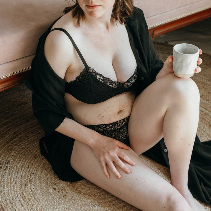 Woman in black lingerie leans against sofa and drinks coffee