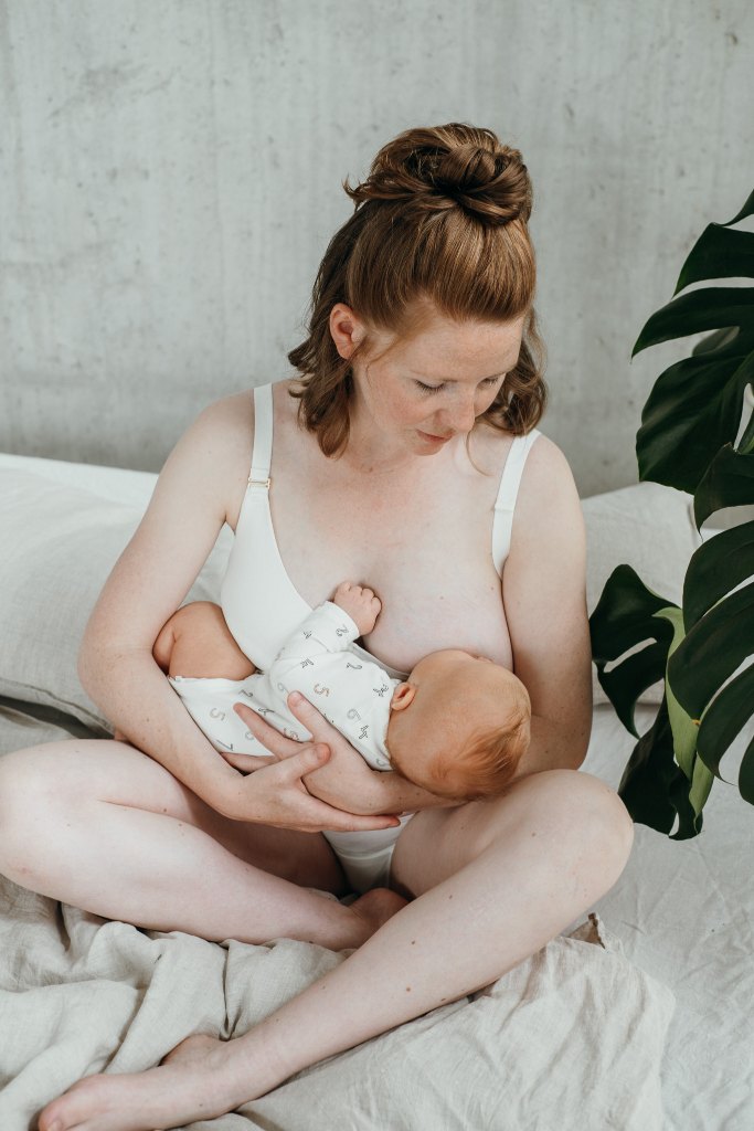 Woman sits on bed and breastfeeds her child. She is wearing a white breastfeedingBralette.