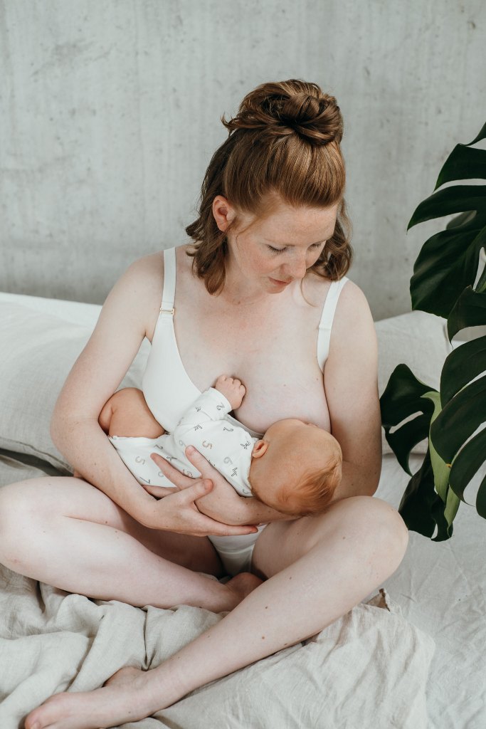 Donna seduta sul letto che allatta il figlio. Indossa un vestito bianco per l'allattamentoBralette.