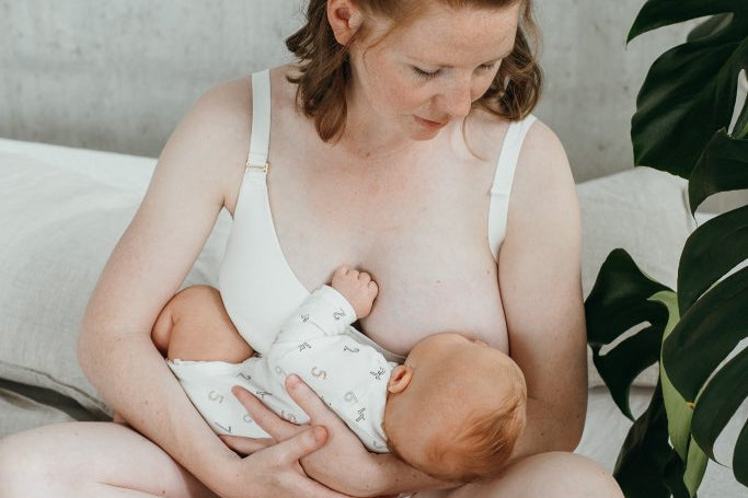 Frau sitzt auf Bett und stillt ihr Kind. Sie Trägt ein weisses Still-Bralette.