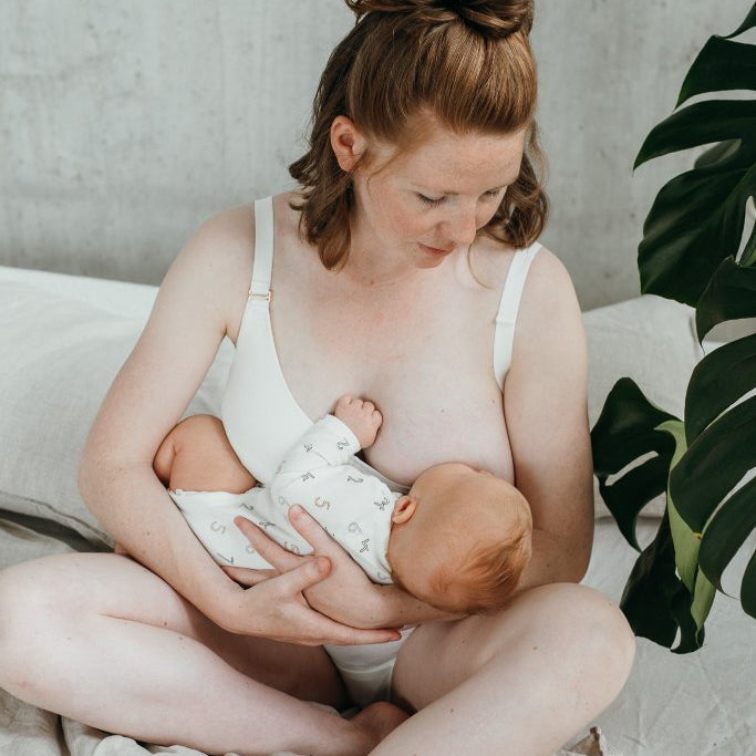 Woman sits on bed and breastfeeds her child. She is wearing a white breastfeedingBralette.