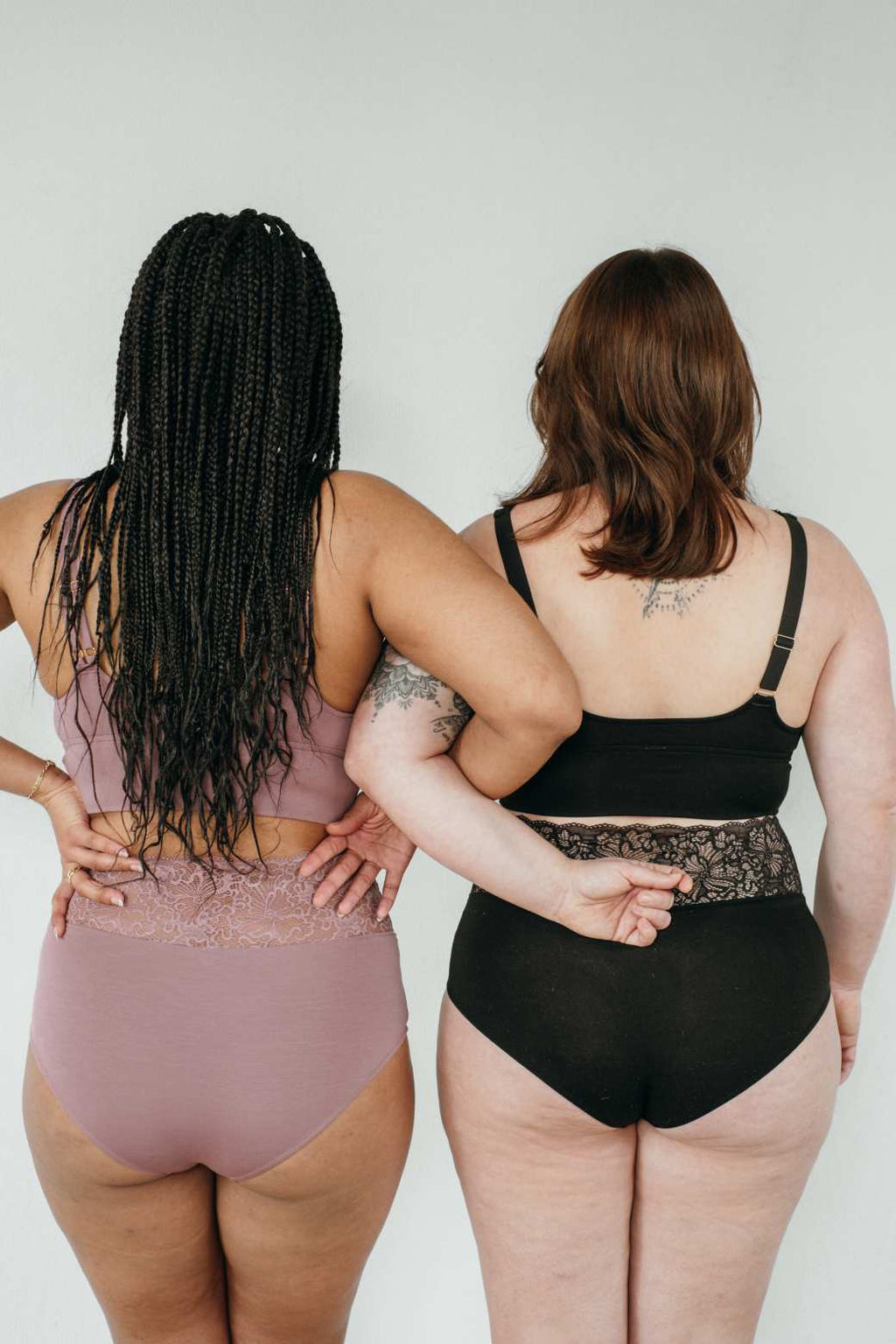 Two women stand in their underwear with their backs to the camera and their arms hooked. 