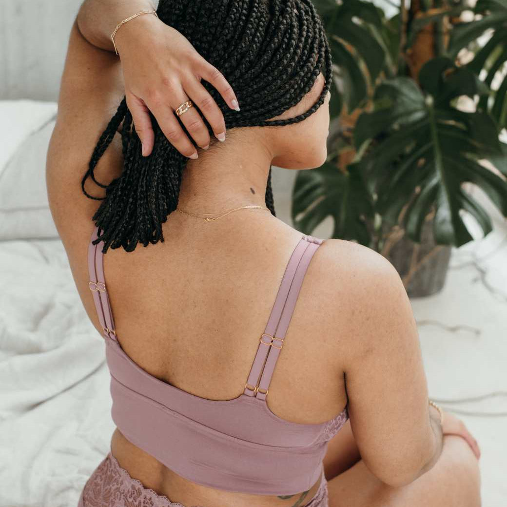 Model in Unterwäsche in Altrosa sitzt auf dem Bett uns streicht die Haare zur Seite.