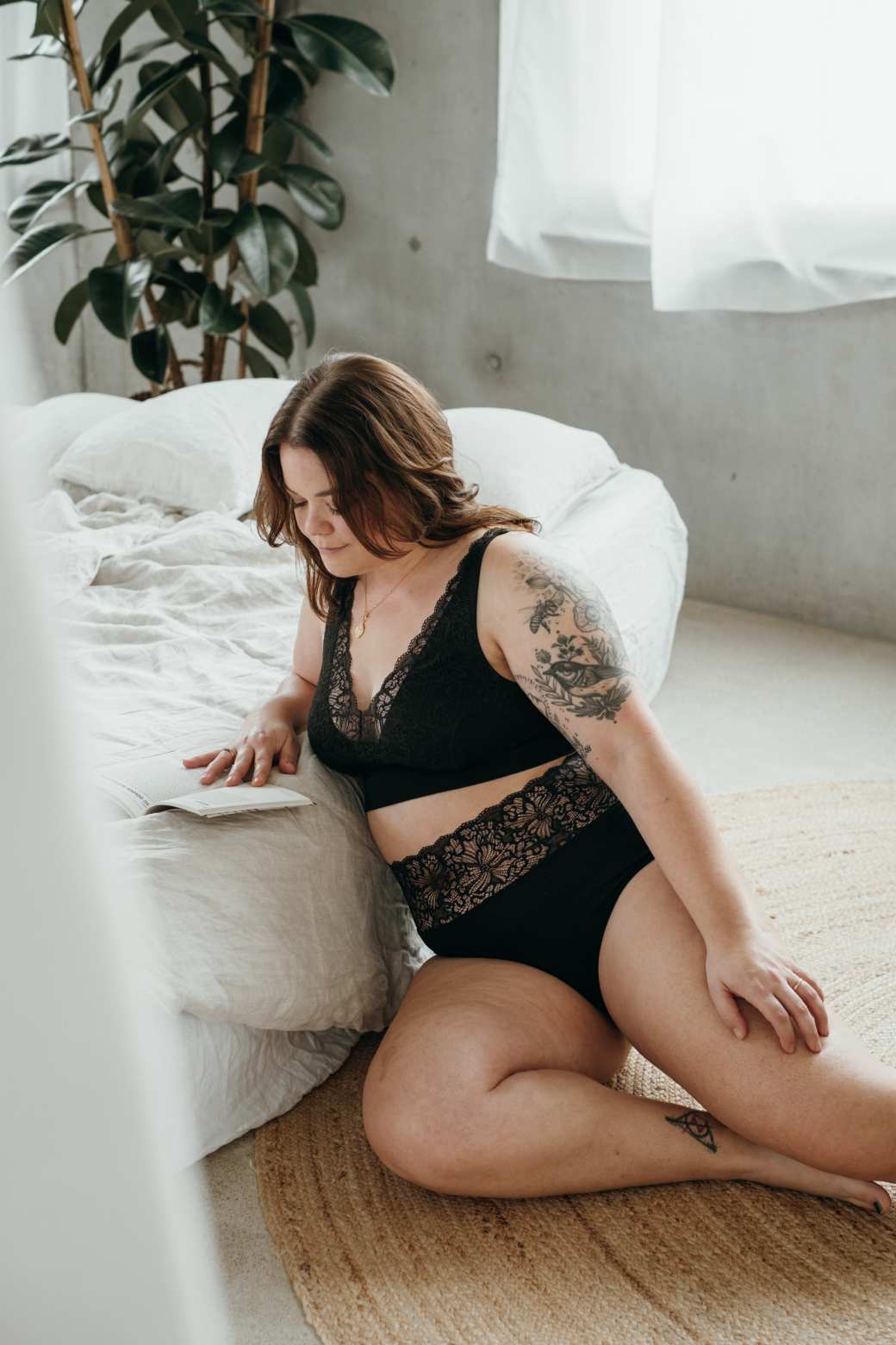 Model in black lingerie made from sustainable materials sits in front of bed and reads.
