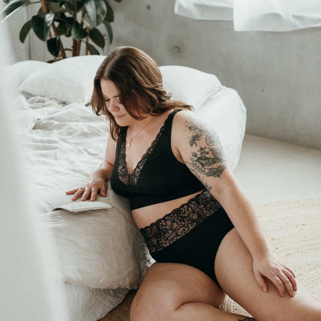 Model in schwarzer Lingerie aus nachhaltigen Materialien sitzt vor Bett und liest.