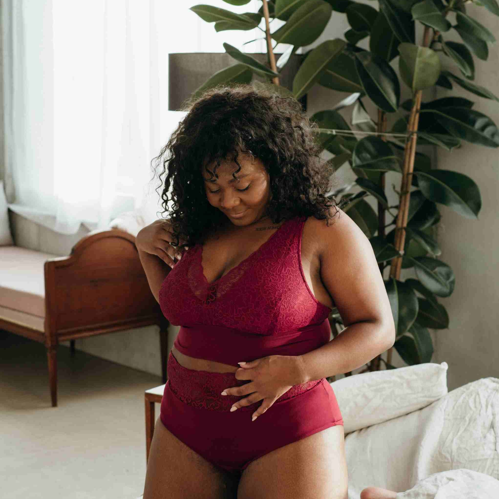 Model in dunkelrotem Dessous aus Spitze und Tencel Lyocell kniet auf Bett.