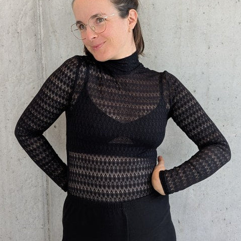 Model trägt den Paula Spitzenpulli und lehnt gegen die Betonwand.