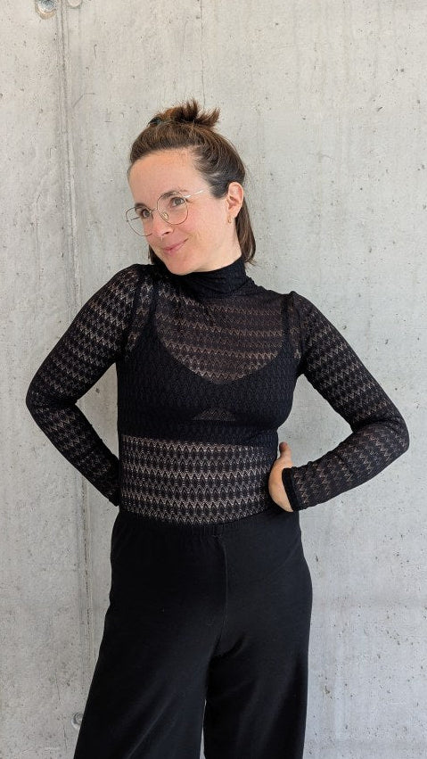 Model trägt den Paula Spitzenpulli und lehnt gegen die Betonwand.