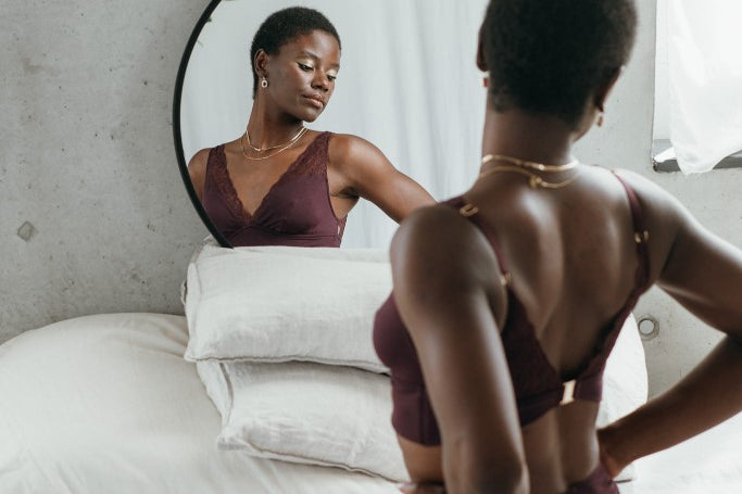 Model kniet auf dem Bett und blickt in den Spiegel. Sie trägt das Sophie Set in der Farbe Aubergine.