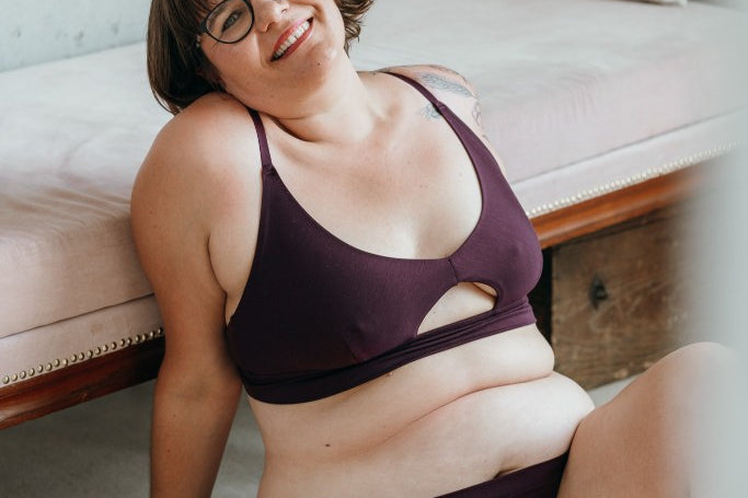 Model trägt das Thea Set in der Farbe Aubergine. Sie sitzt auf dem Boden und lehnt sich an ein Tagesbett an. Sie hält den Kopf leicht geneigt und lächelt in die Kamera.