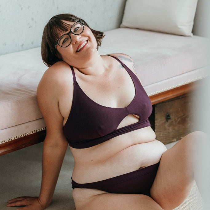 Model trägt das Thea Set in der Farbe Aubergine. Sie sitzt auf dem Boden und lehnt sich an ein Tagesbett an. Sie hält den Kopf leicht geneigt und lächelt in die Kamera.