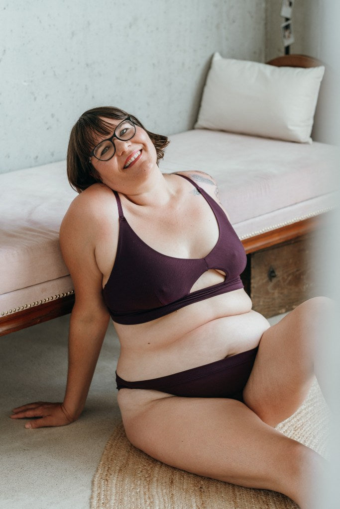 Model trägt das Thea Set in der Farbe Aubergine. Sie sitzt auf dem Boden und lehnt sich an ein Tagesbett an. Sie hält den Kopf leicht geneigt und lächelt in die Kamera.