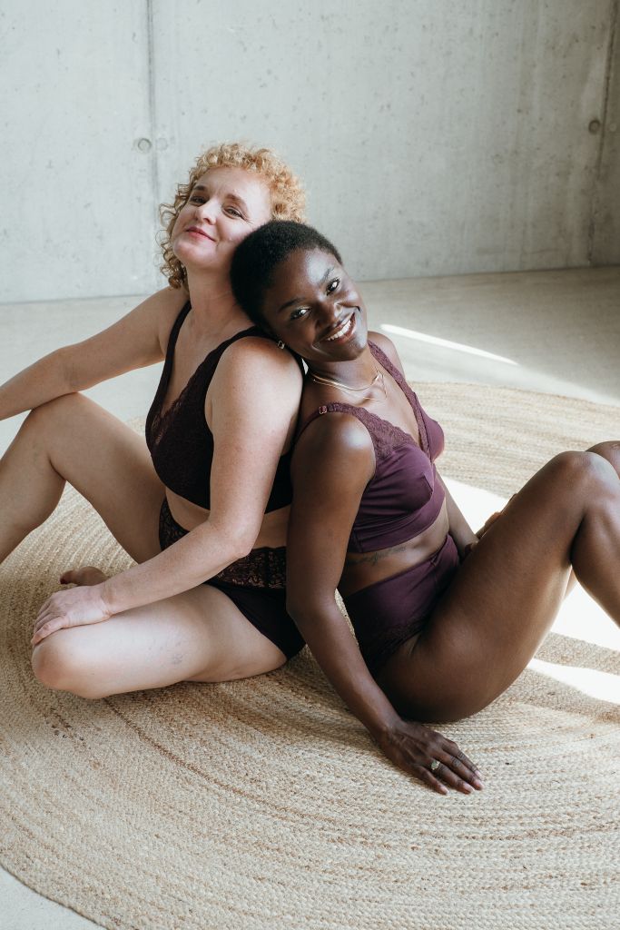 Zwei Models sitzen Rücken an Rücken auf dem Boden und lächeln in die Kamera. Die eine trägt das Nelly Set und die andere das Sophie Set in der Farbe Aubergine.