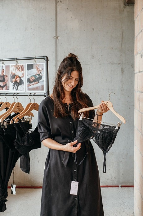 Foto zeigt Kira. Sie steht an einer Kleiderstange und hält ein Bralette am Kleiderbügel hoch.