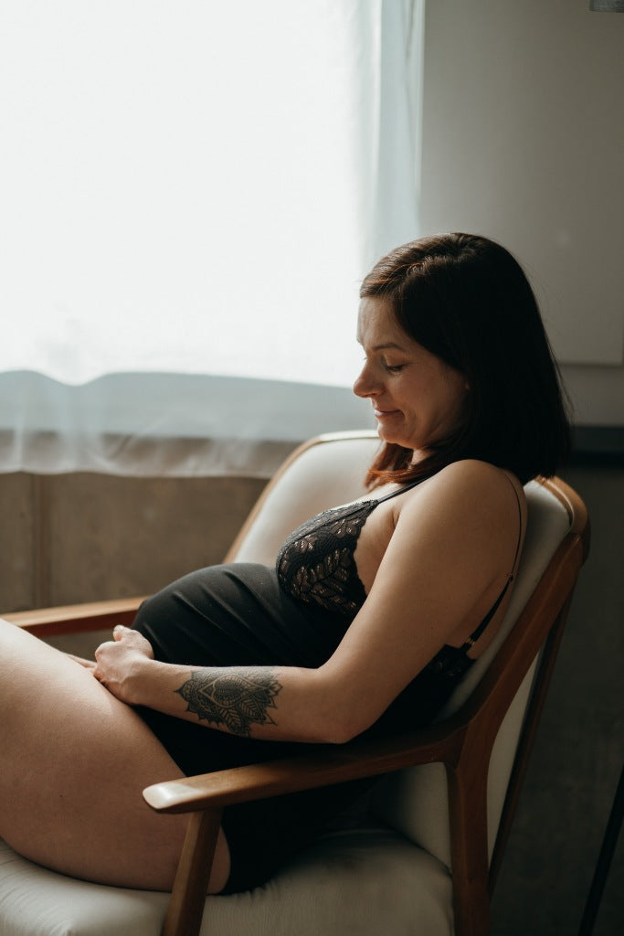 Woman with baby bump wears playful lace body Jina and sits on white armchair.