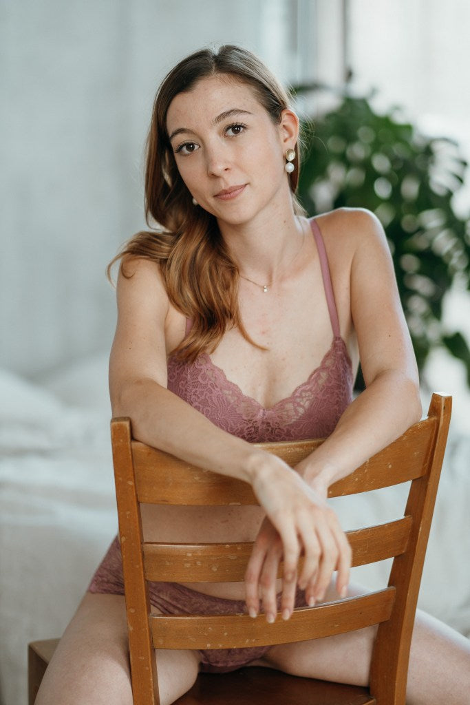Model in dusky pink lace bralette with removable pads sits upside down on chair.
