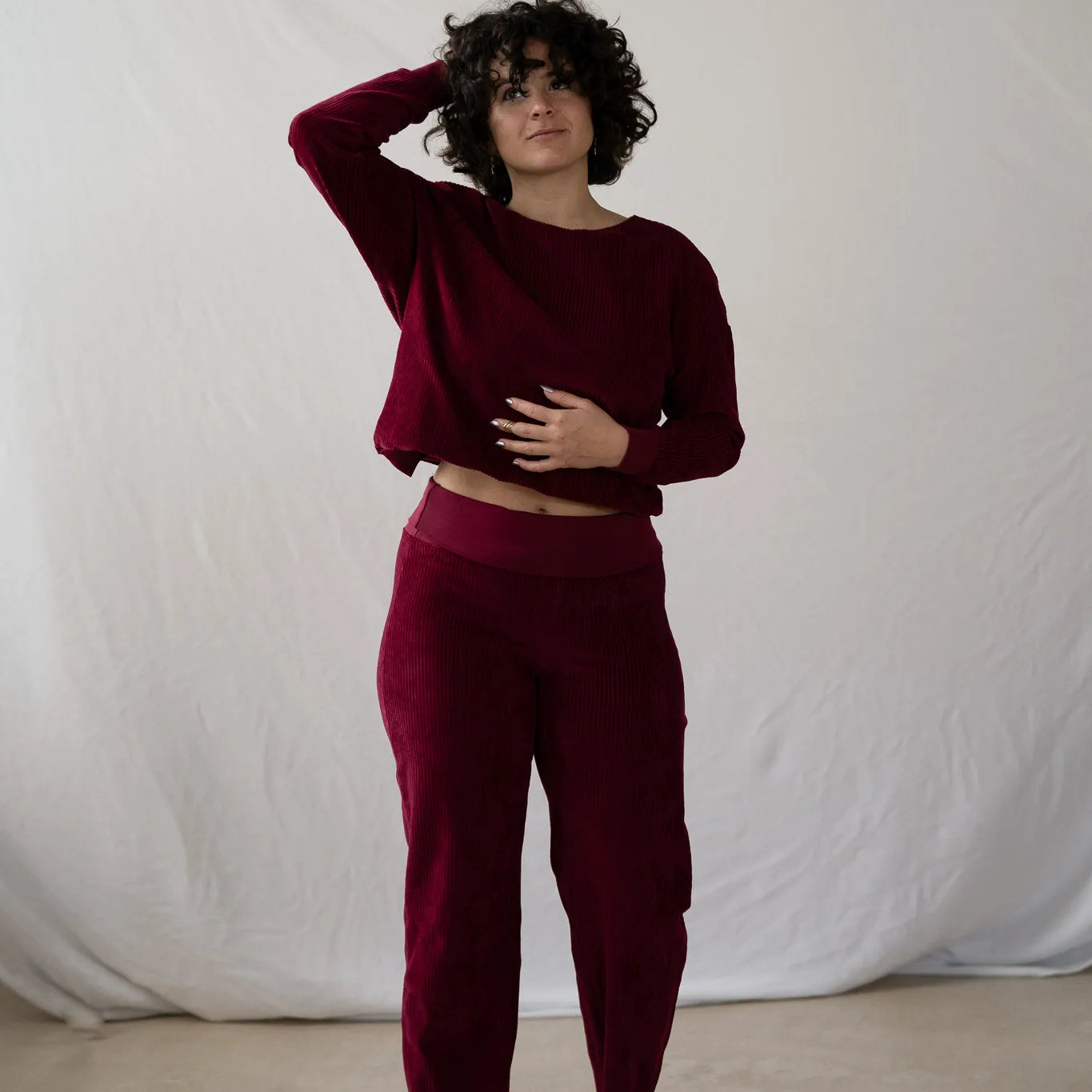 Model steht vor weissem Hintergrund, sie trägt den Chloé Pullover und die Kelly Hose in Ruby Red. Mit einer Hand wuschelt sie durch ihr lockiges kurzes Haar.