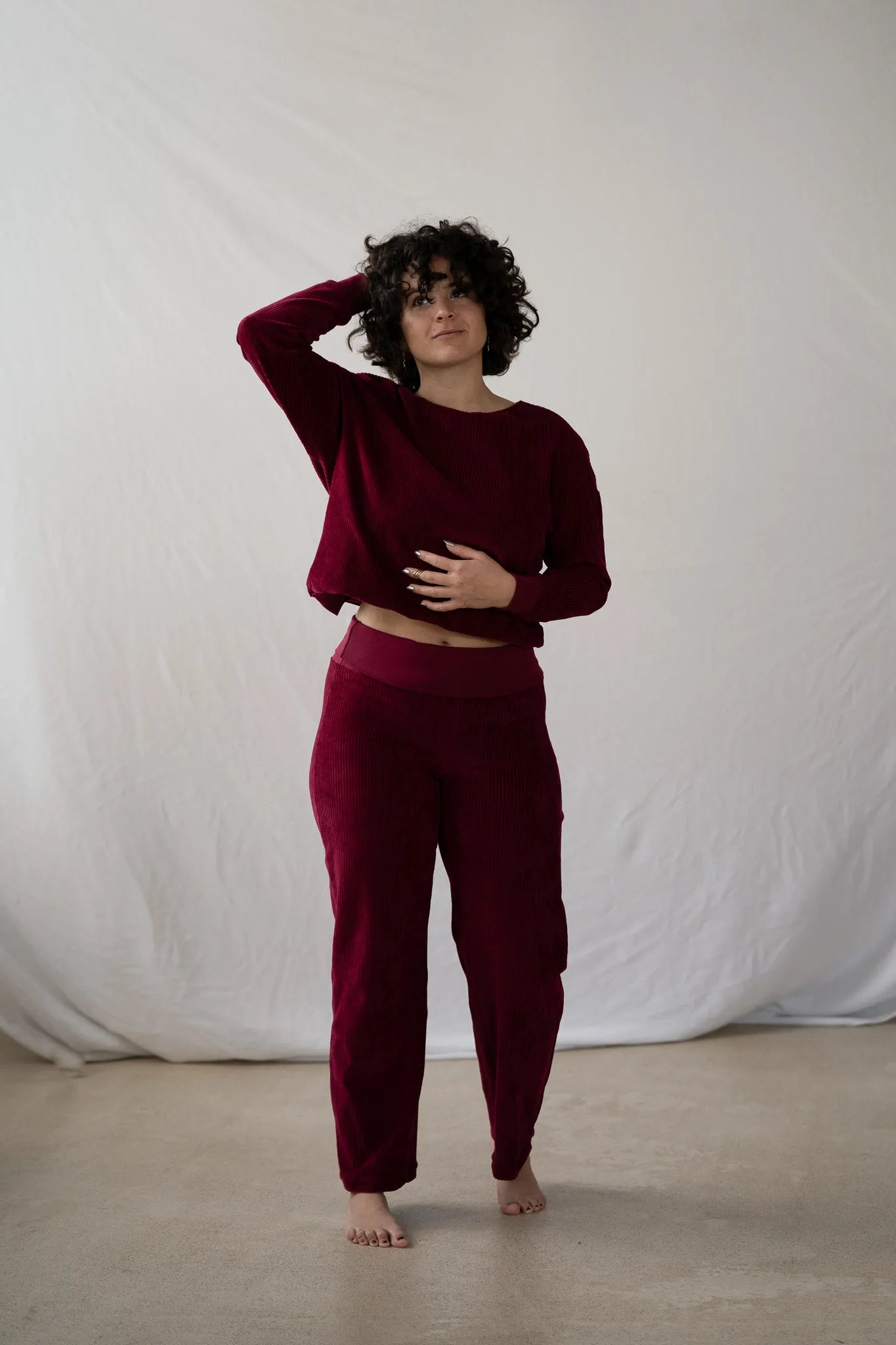 Model steht vor weissem Hintergrund, sie trägt den Chloé Pullover und die Kelly Hose in Ruby Red. Mit einer Hand wuschelt sie durch ihr lockiges kurzes Haar.